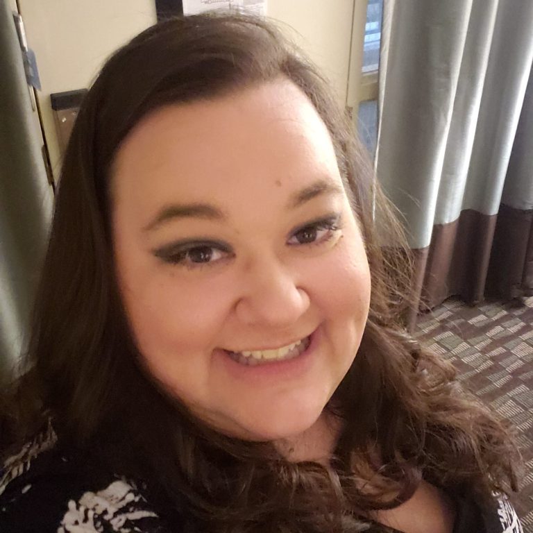 Smiling person with long hair, wearing a black and white patterned top, indoors.