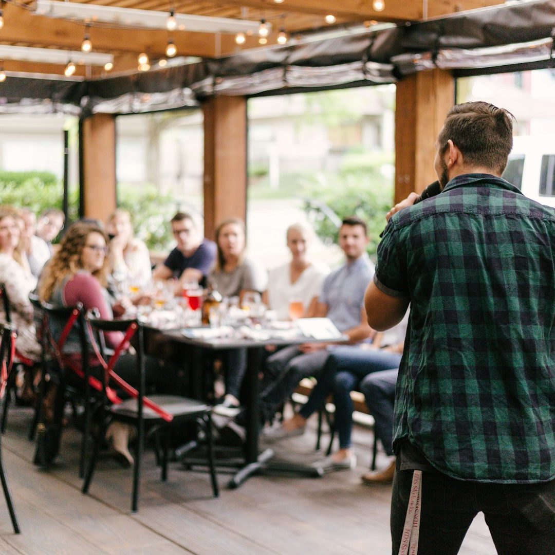 Community Event, Man Speaking A man speaks to a group of people gathered at a restaurant table outdoors.