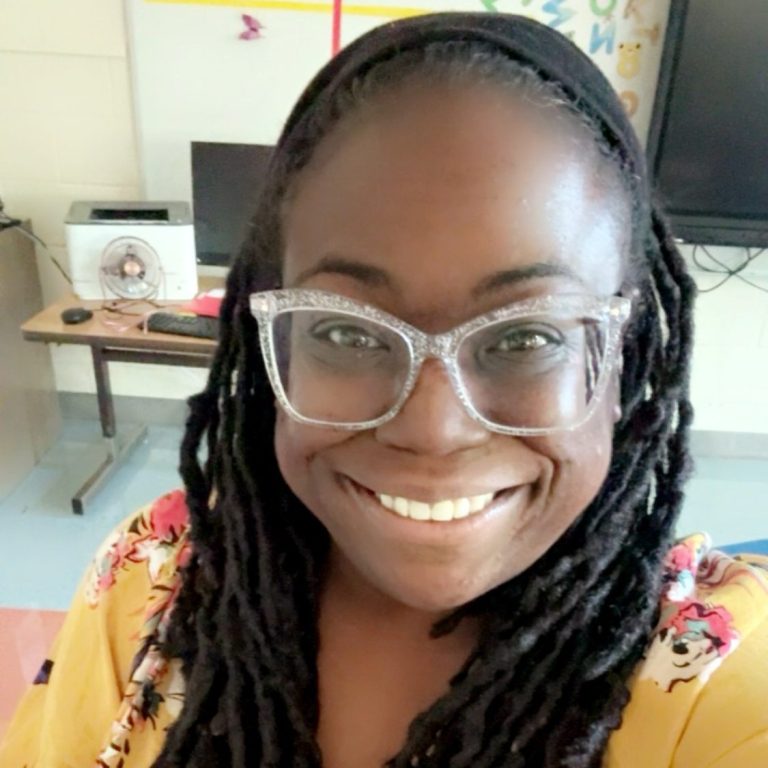 Smiling woman with glasses and long hair, wearing a colorful top, in a classroom setting.
